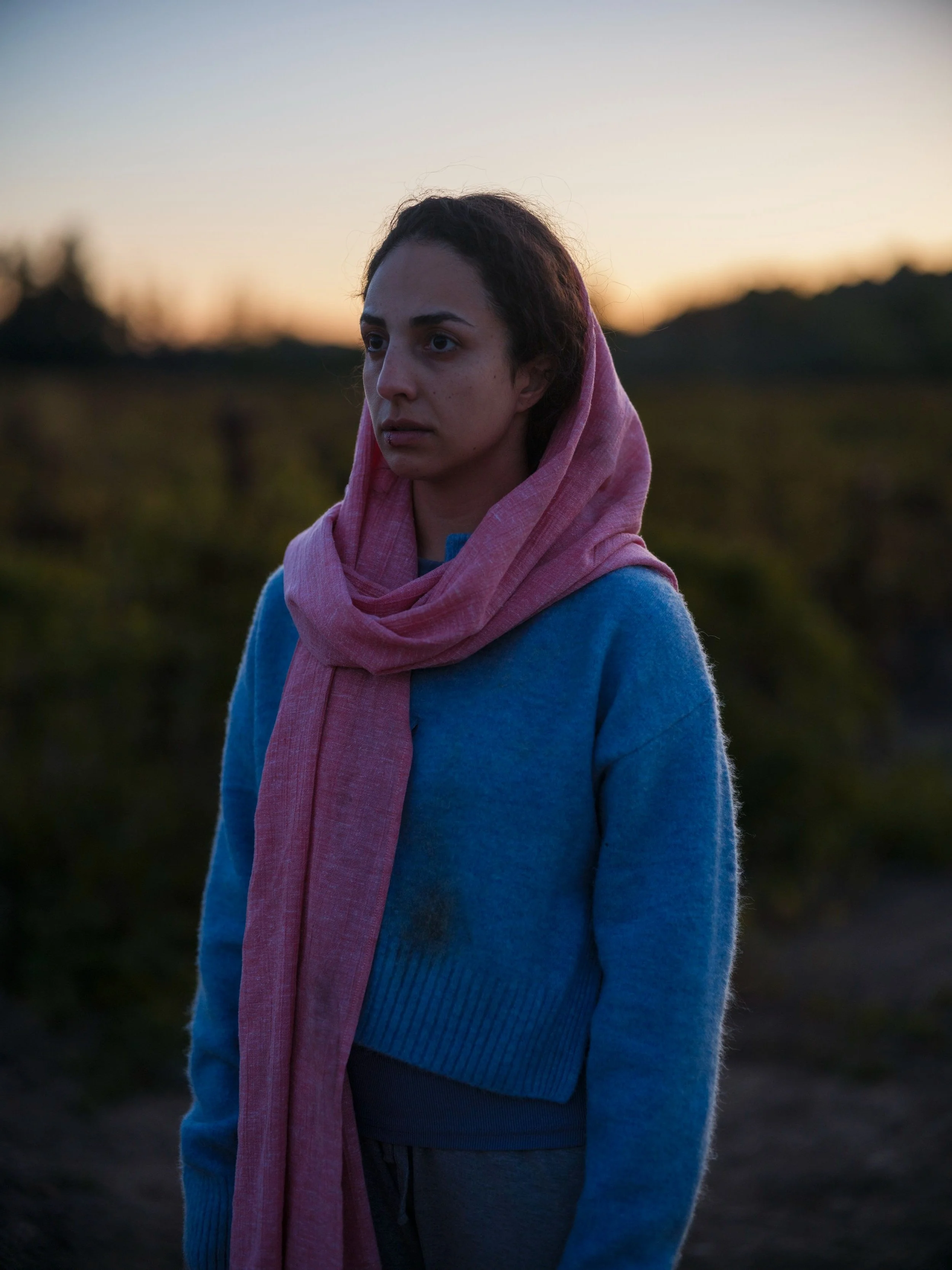 A woman with a pink scarf wrapped around her head and a blue sweater standing outdoors at sunset, with a blurred background of nature.