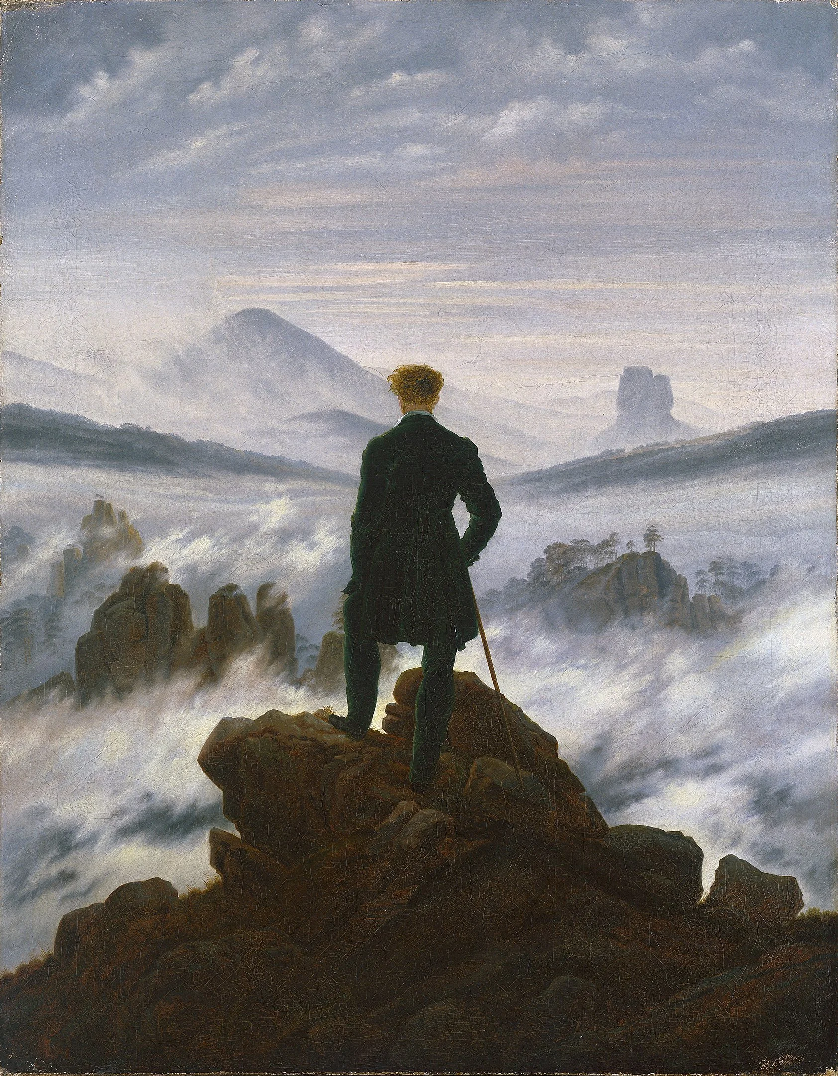 Un uomo in abito nero si trova su una roccia, guarda un paesaggio montuoso con nuvole e nebbia, alcuni picchi si distinguono sullo sfondo.