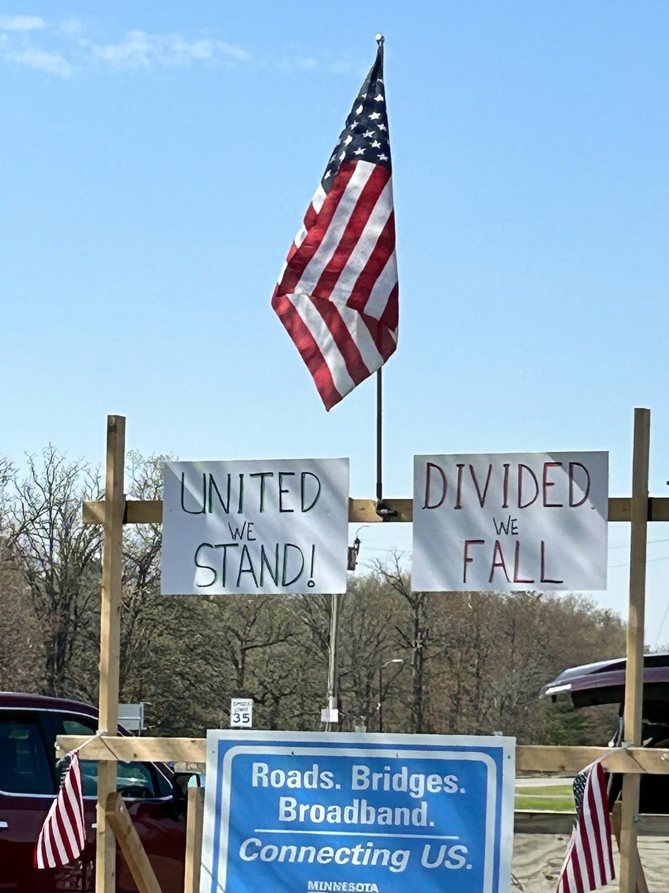 A political protest display with an American flag and signs that read: 'United we stand!', 'Divided we fall'. The display is on a trailer constructed with wooden frames and decorated with American flags. Our Aitkin Community comes first!