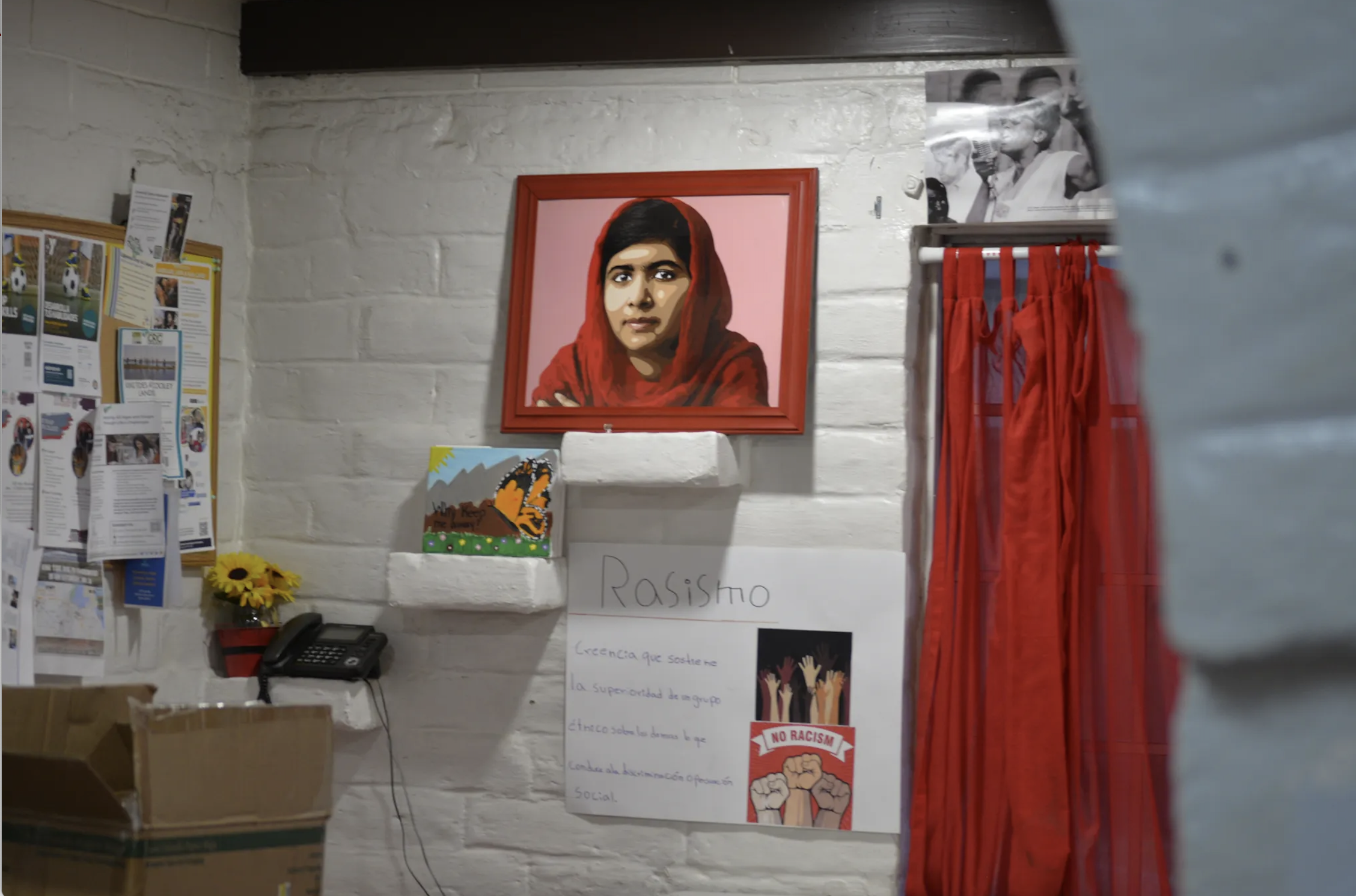 A mural of a woman wearing a red headscarf hangs on a white brick wall inside a room. To the left, there are bulletin boards with papers and flyers. A black telephone and a sunflower in a red pot are on a white shelf below the mural. Next to the shelf is a white poster with the word 'Rasísmo' and colorful raised hands with a 'No Racism' graphic. There are red curtains hanging on a rod to the right of the mural.