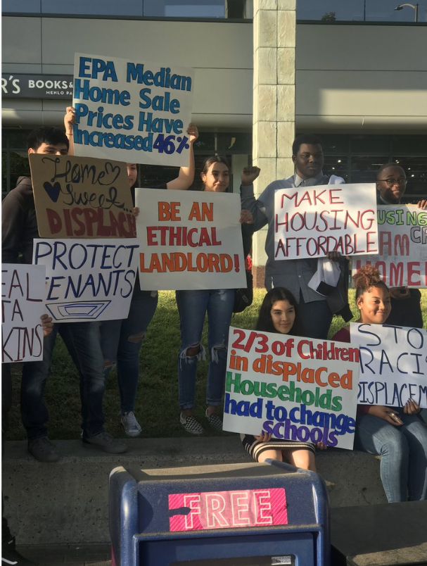 Group of young people protesting, holding signs about housing, displacement, and ethics outside a building with a sign on a bus stop that reads 'FREE'.