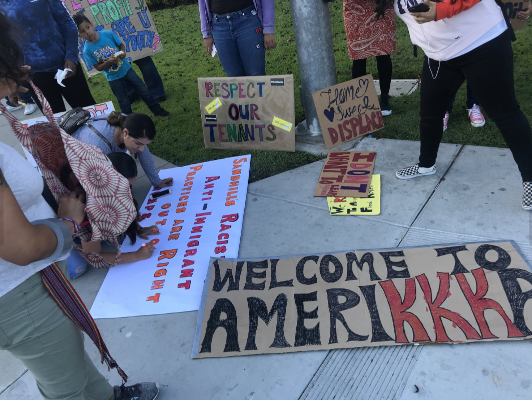 People holding signs and banners at a protest or demonstration, including a large sign that reads 'Welcome to America' and another that says 'Racists, Anti-American practices are your right'.
