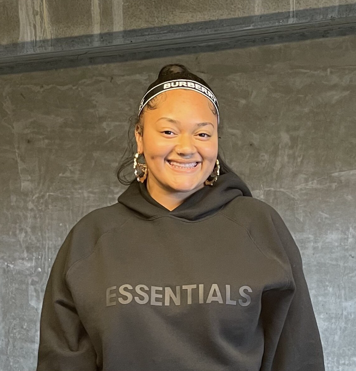 A smiling woman wearing a black hooded sweatshirt with the word 'ESSENTIALS' printed on it and a black headband with white lettering, standing against a textured gray wall.