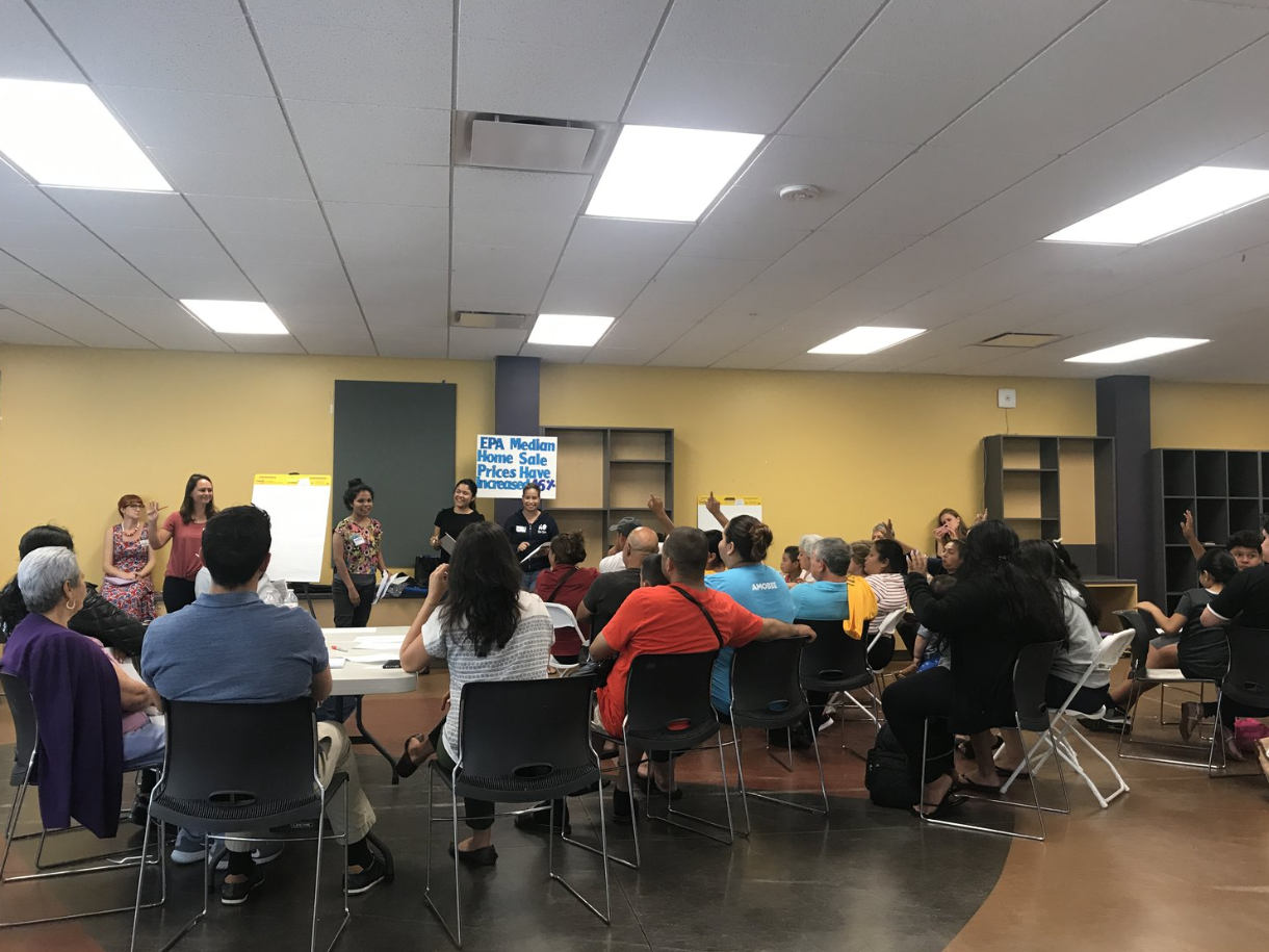 A diverse group of people sitting around tables in a conference room, attending a presentation. Several individuals are raising their hands. Four women are standing at the front near a yellow presentation board, and a digital sign on the wall reads '