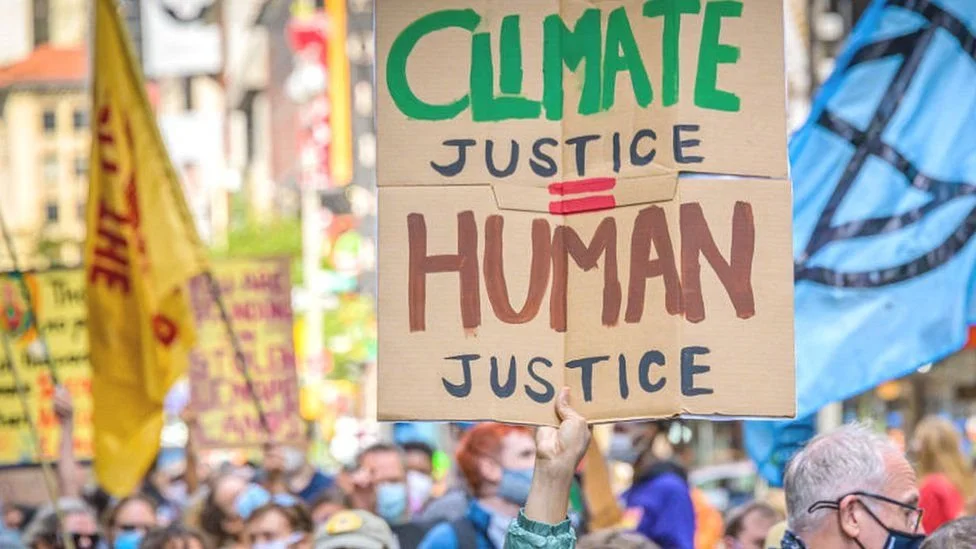 A protester holding a cardboard sign that reads 'Climate Justice = Human Justice' during a crowded demonstration.
