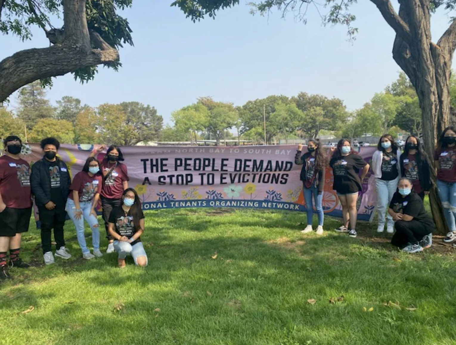 Group of people holding a large pink banner that reads 'The People Demand a Stop to Evictions' at an outdoor park with trees and green grass, all wearing masks.