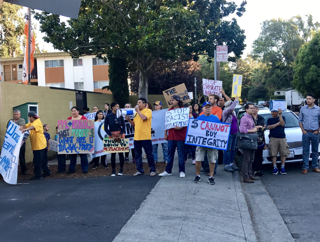 Group of people protesting on a sidewalk holding signs with messages about ethics, corporate landowners, and displacement.