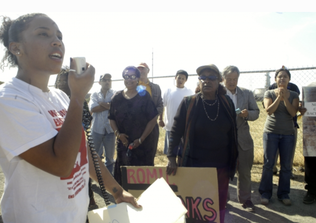 A woman speaking into a microphone at a protest or rally, with a group of people listening and holding signs behind her.