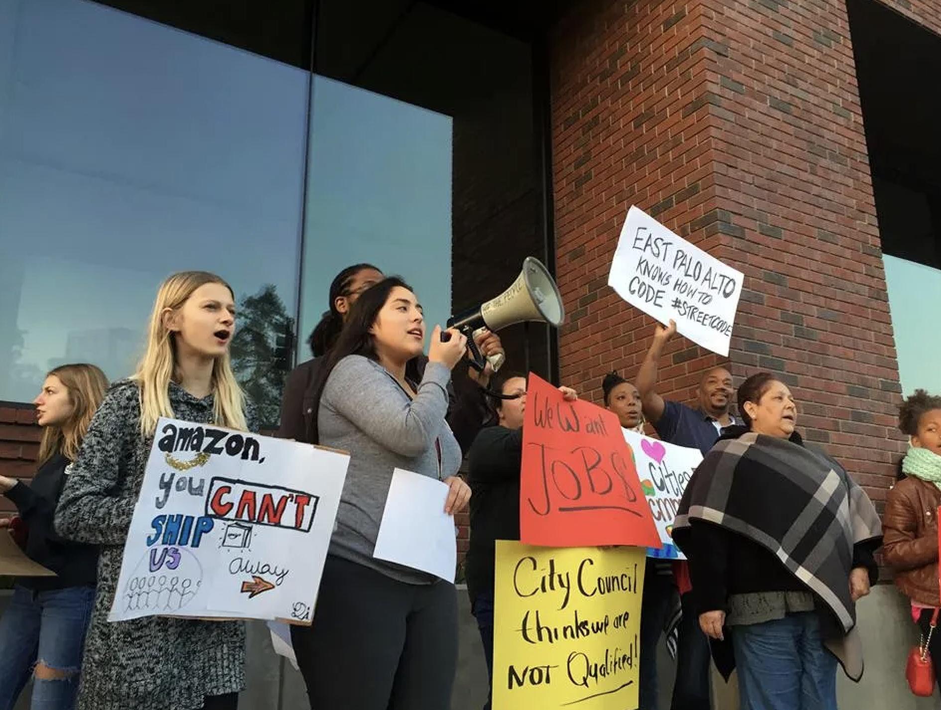 Group of protesters holding signs during a demonstration outside a building with brick and glass facade. Signs include messages about Amazon, jobs, and city council.