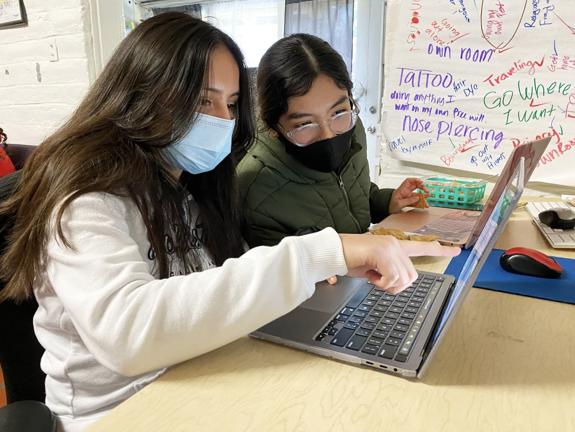 Two women wearing masks sit at a table working on laptops. One points at her laptop screen, and a colorful wall with handwritten notes and drawings is in the background.