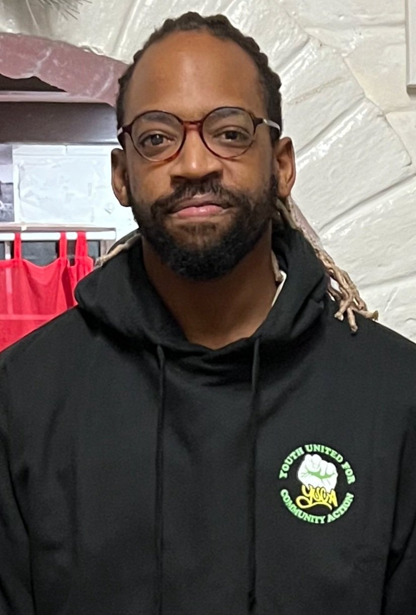 A man with glasses, a beard, and dreadlocks wearing a black hoodie with a logo that says Youth United for Community Action, standing indoors with a white wall and some red clothing in the background.