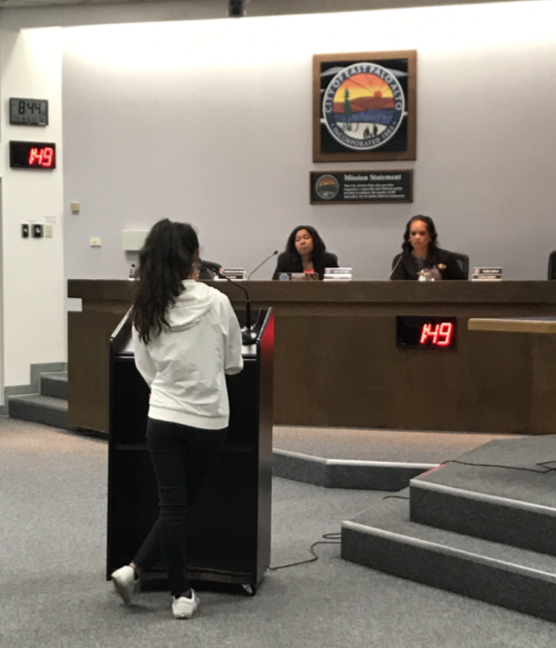 A young woman in a white hoodie and black pants standing at a podium speaking to a panel of three women seated at a long desk in a government or city council chamber. The room has a clock and a digital display showing the number 49, and a sunset and 