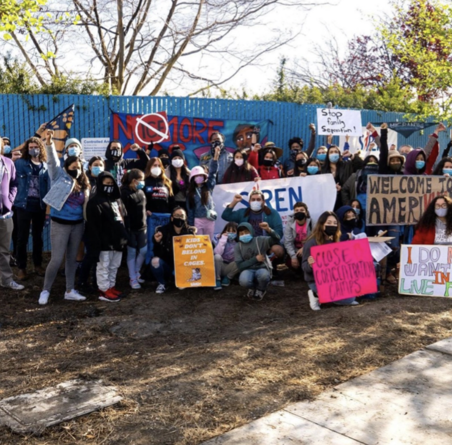 Group of people protesting outdoors, holding signs with messages like "Stop family Separation!", "Welcome to America!", and "Close Concentration Camps"; group wearing masks, some raising fists, in front of a blue fence with colorful graffiti, trees i