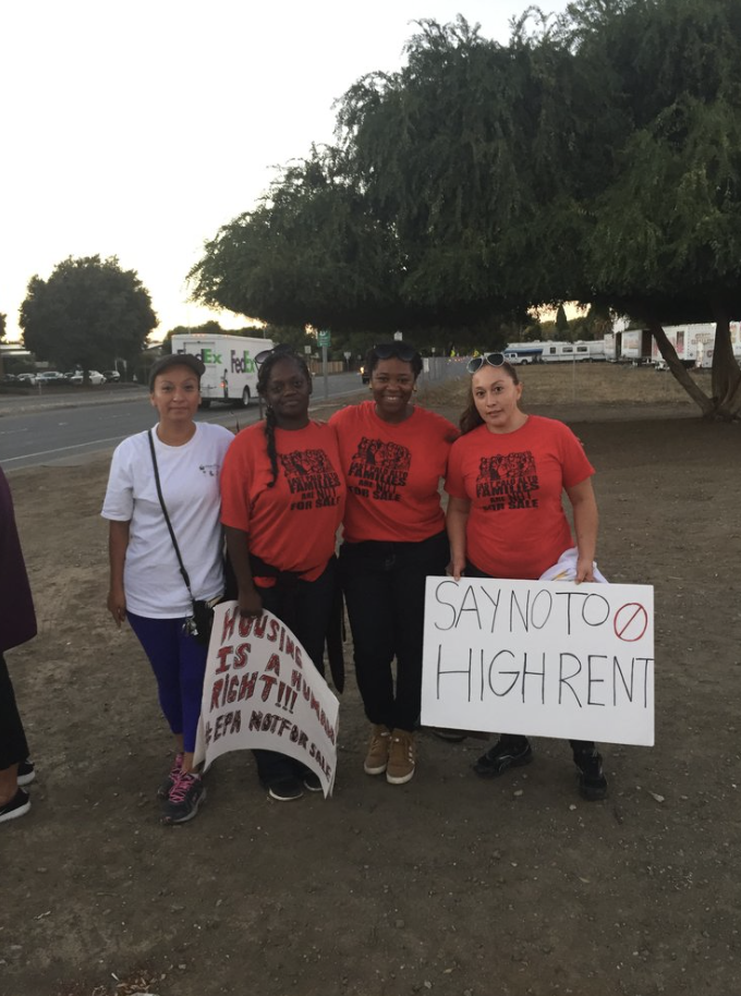 Four women standing outdoors with trees and a road in the background, holding protest signs about housing issues.