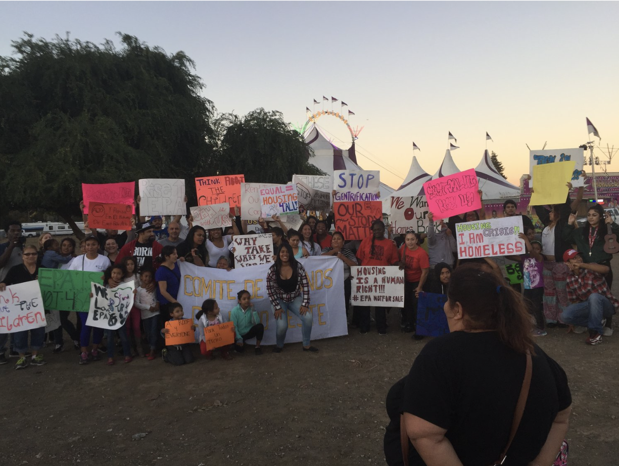 A large group of people, including children and adults, gathered at an outdoor protest or rally. They are holding various signs advocating for housing rights, fighting gentrification, and ending homelessness. The background shows carnival tents and a
