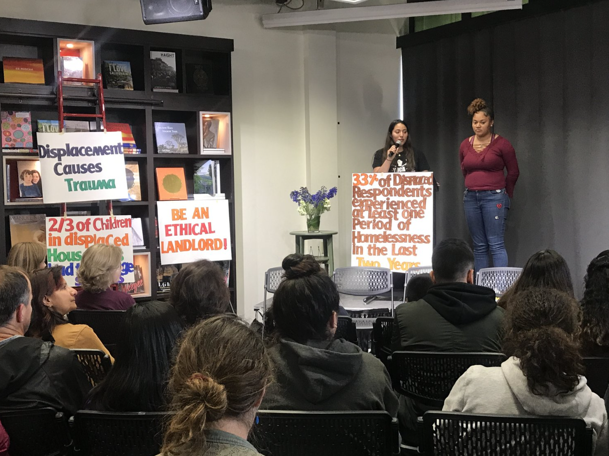 A woman standing on a stage speaking into a microphone during a presentation, with another woman standing beside her. The audience is seated and listening. Three large informational posters with facts about displacement, trauma, and homelessness are 