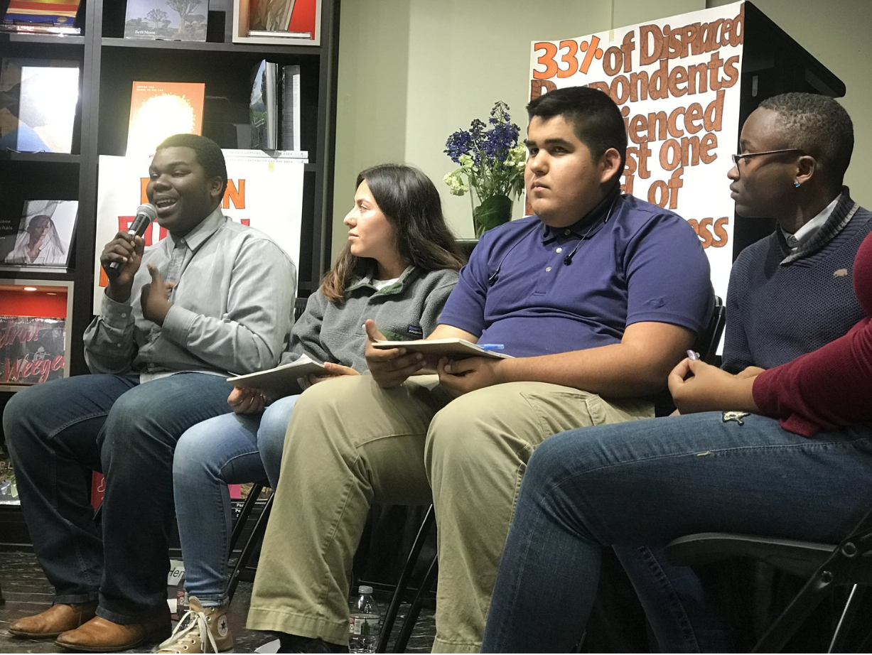 Young man with a microphone speaking during a panel discussion, seated with three other young adults, in a room with bookshelves and a poster about disinformation in the background.