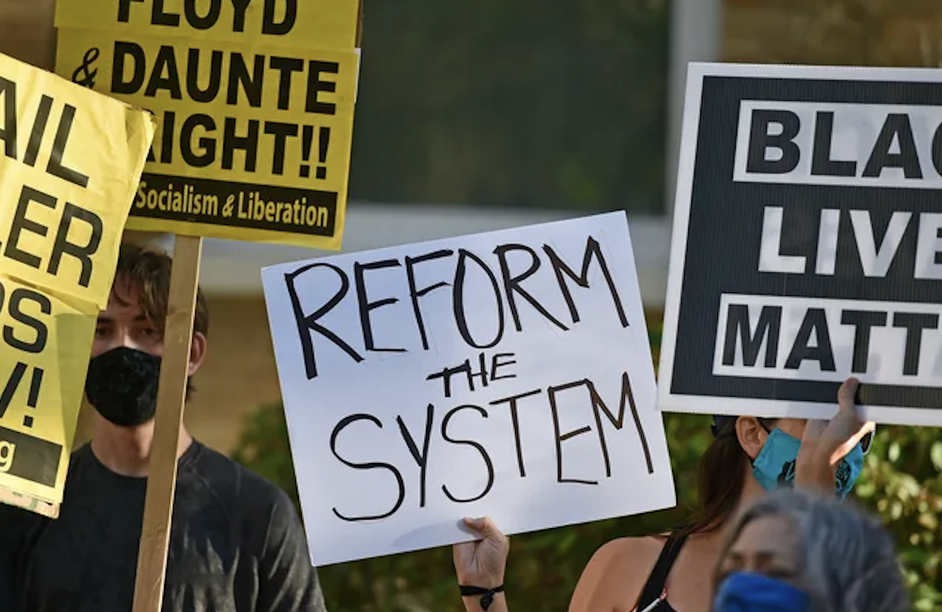 Protesters holding signs that say 'Reform the System' and 'Black Lives Matter' at a rally.