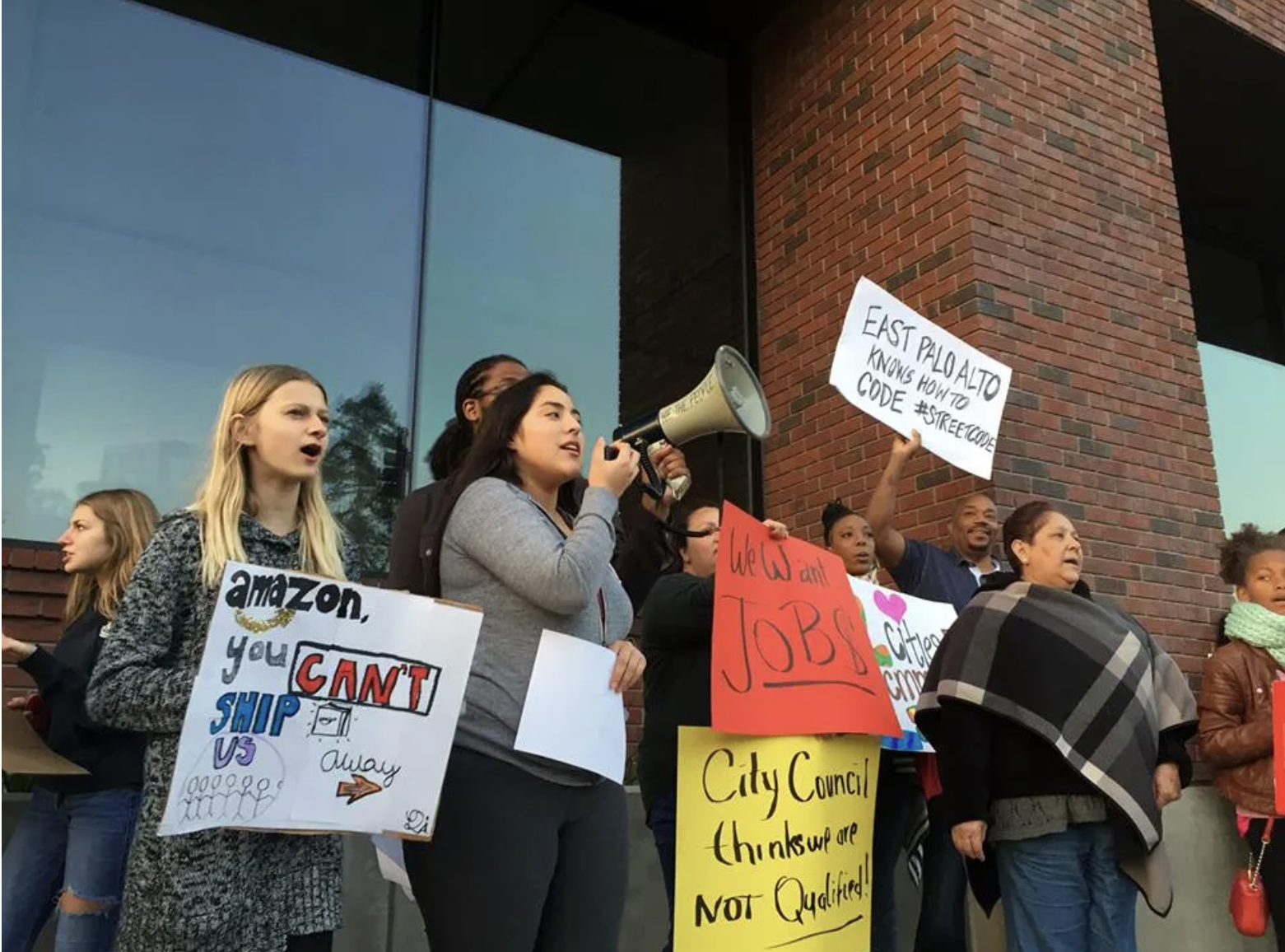 Group of diverse protesters holding signs and speaking out during a rally against job and immigration issues outside a building with reflective windows and brick wall.
