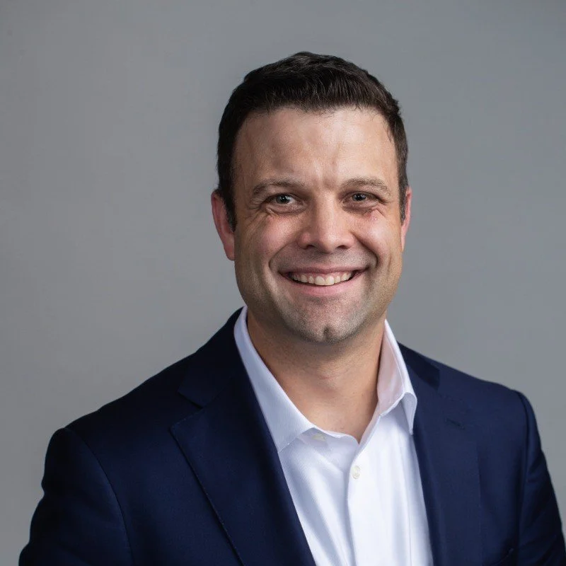 A smiling man in a navy suit and white shirt against a gray background.