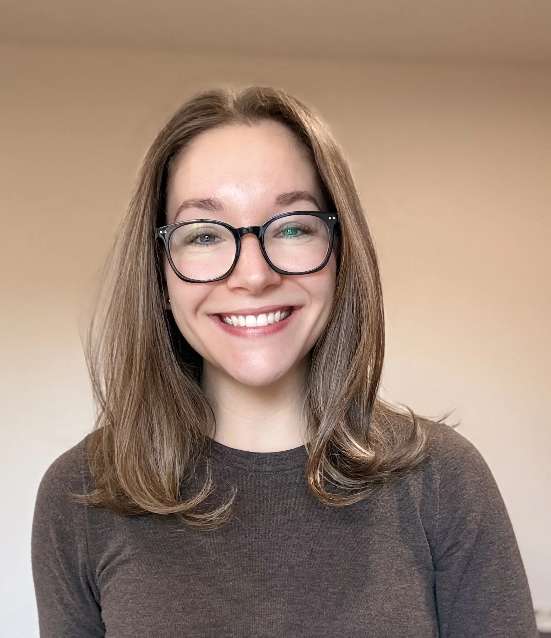 Close-up of a woman with long, light brown hair, wearing large black glasses and a dark brown sweater, smiling against a neutral beige background.