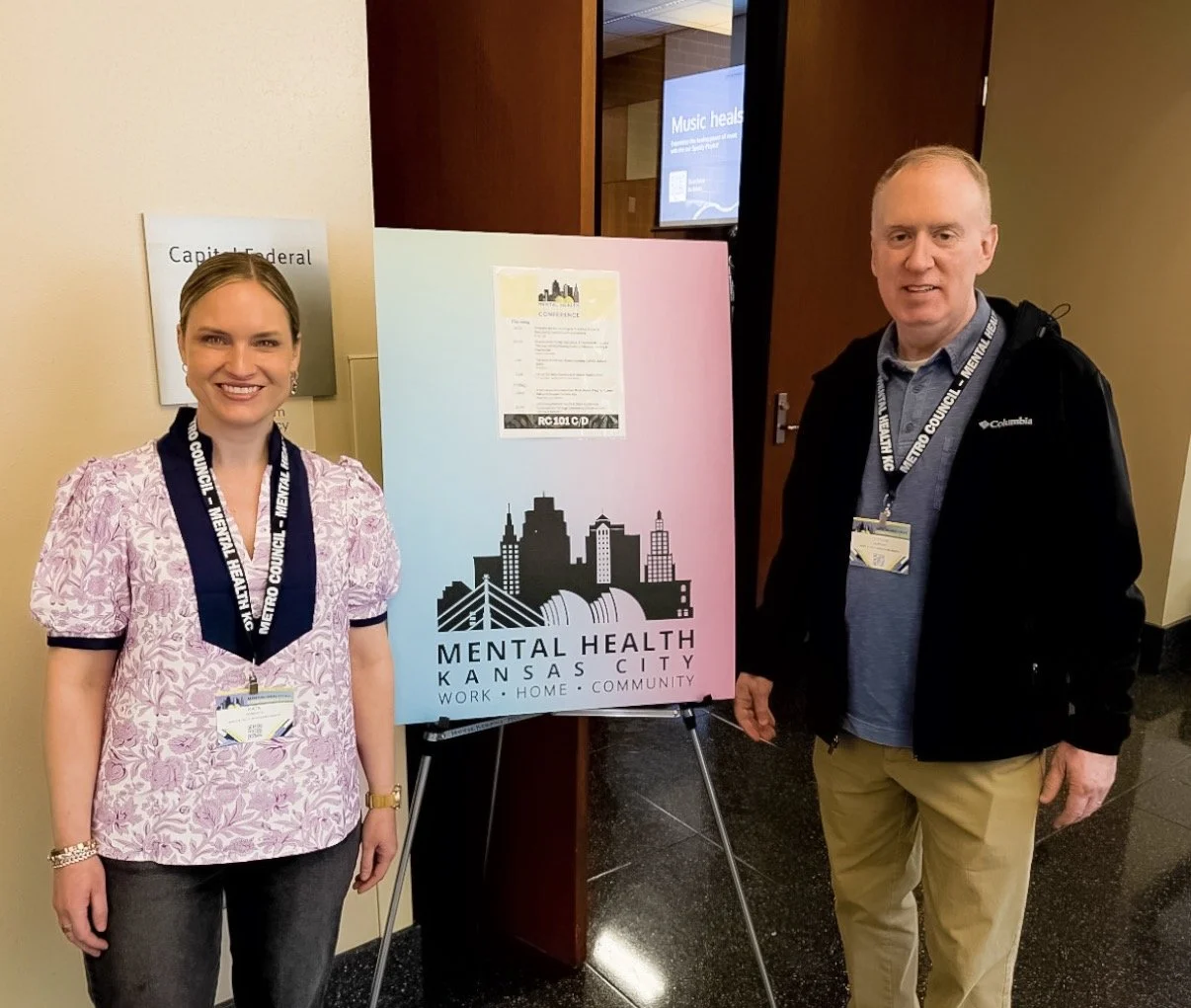 Kate Horowitz, LSCSW, and Steve Murdock, LCPC, of Mercy & Truth's behavioral health team smile beside the Mental Health Kansas City "Work · Home · Community" conference sign at the 2026 Mental Health KC Conference.