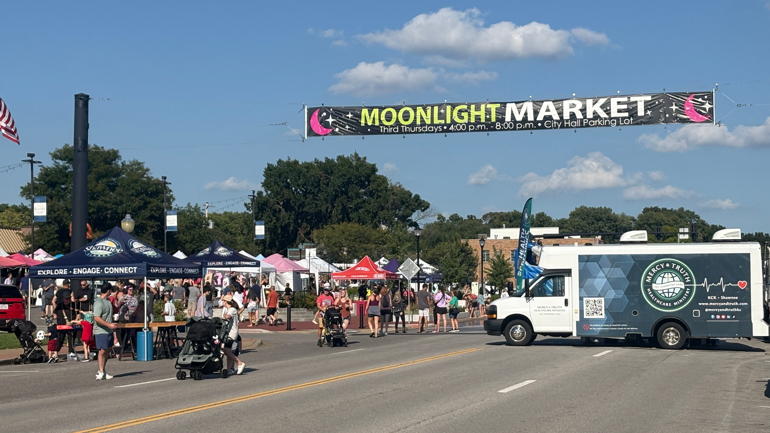 Mercy & Truth Healthcare Ministry's mobile health clinic parked along Johnson Drive during Moonlight Market in downtown Shawnee, Kansas, as families walk among vendor tents beneath the market banner.