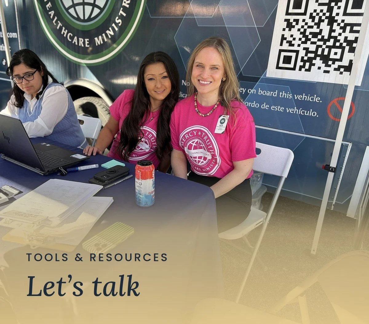 Smiling Mercy & Truth team members in pink shirts welcome guests at the Mobile Health Clinic sign-in table. Text reads "Tools & Resources: Let's talk."