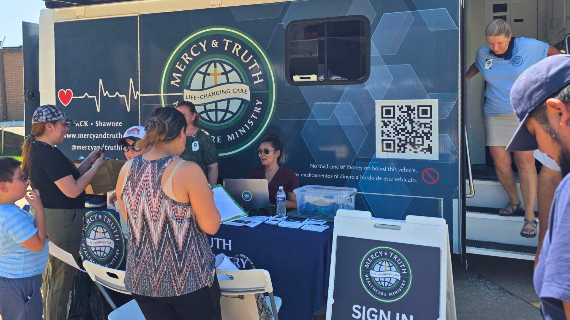 Visitors sign in at the Mercy & Truth mobile health clinic during a free Wellness Hub event in Shawnee, Kansas, as staff greet families at the table.