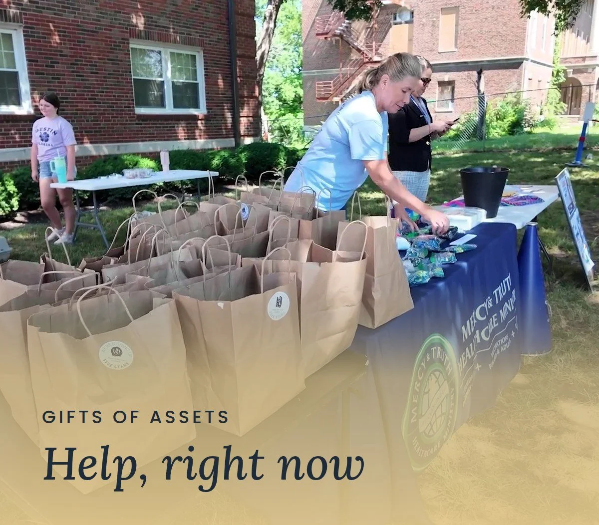 Volunteers prepare rows of free grocery bags at a Mercy & Truth outreach table. Text reads "Gifts of Assets: Help, right now."