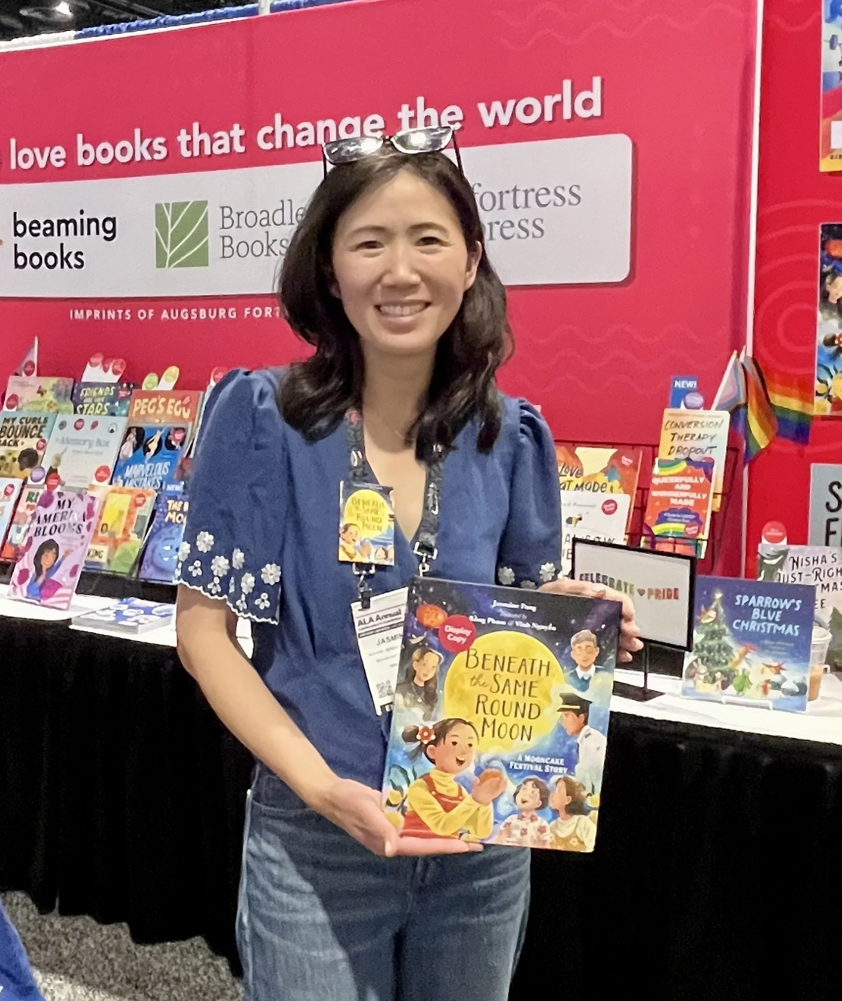 A woman smiling at a book fair holding a children's book titled 'Beneath the Same Round Moon'. She wears a blue shirt with floral embroidery and has glasses resting on her head. The background features a red display with books and signs about love books and relevance to social causes.