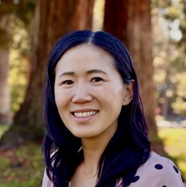 A woman with dark blue hair smiling outdoors with trees in the background.