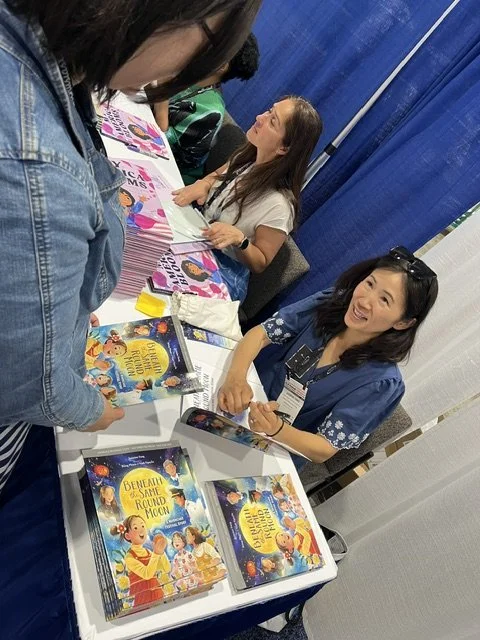 A woman and a young girl are sitting at a book table, smiling and engaging with visitors at a book fair. The table displays children's books, including 'Demonese and the Small Round Food.' The girl is talking to a visitor.