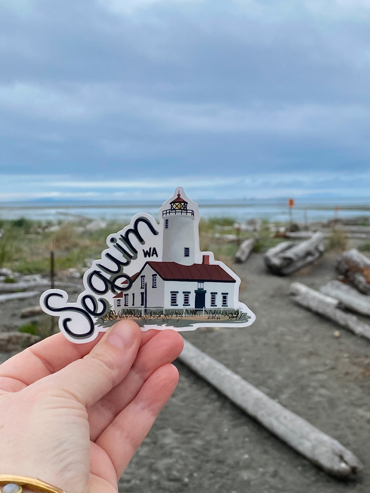 Hand holding a sticker with an illustration of a lighthouse and the words 'Seautim WA' against a beach and cloudy sky background.