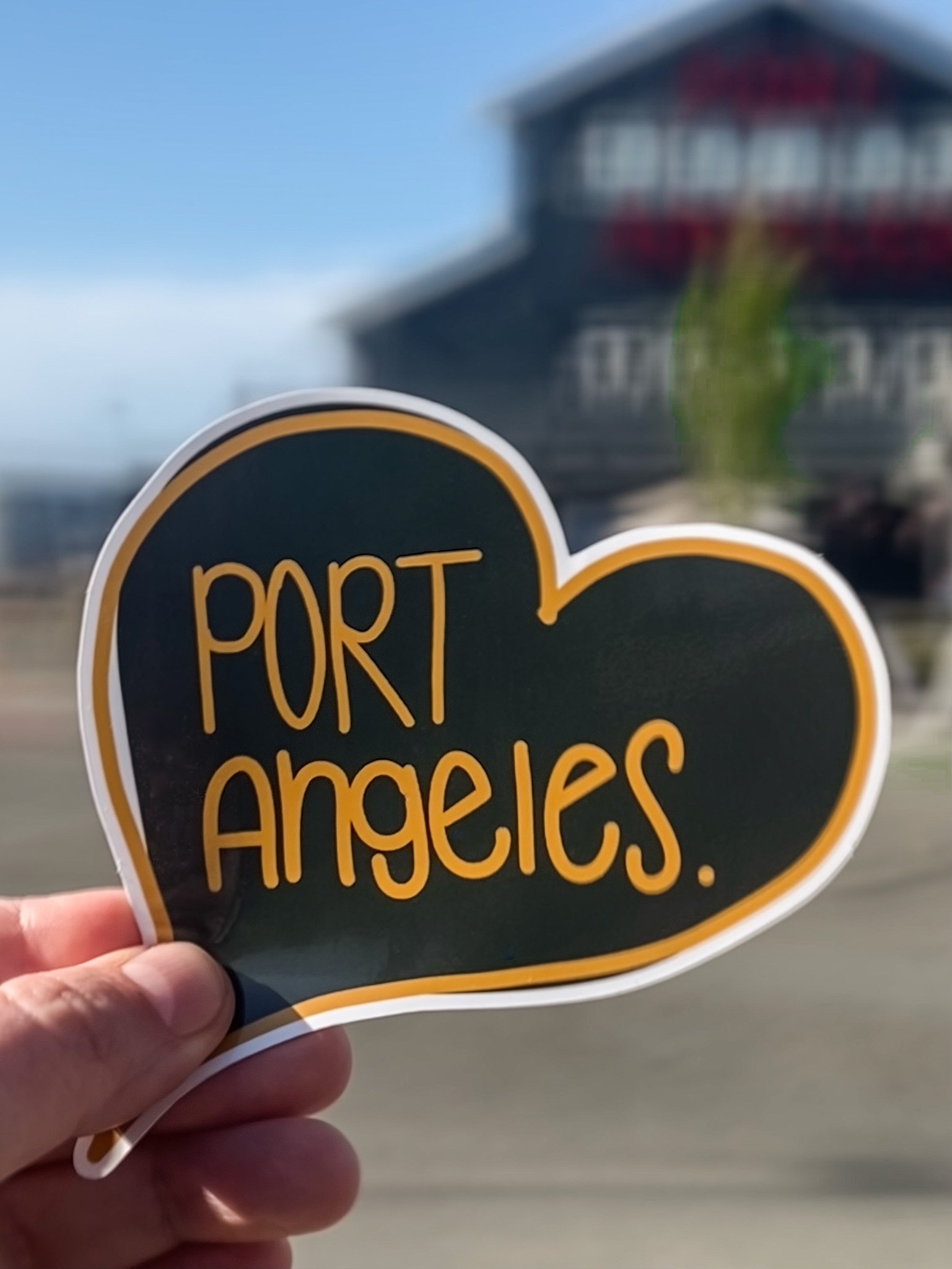 A hand is holding a sticker that says "PORT ANGELES" in orange letters on a dark background with a yellow border, with a blurred building in the background.