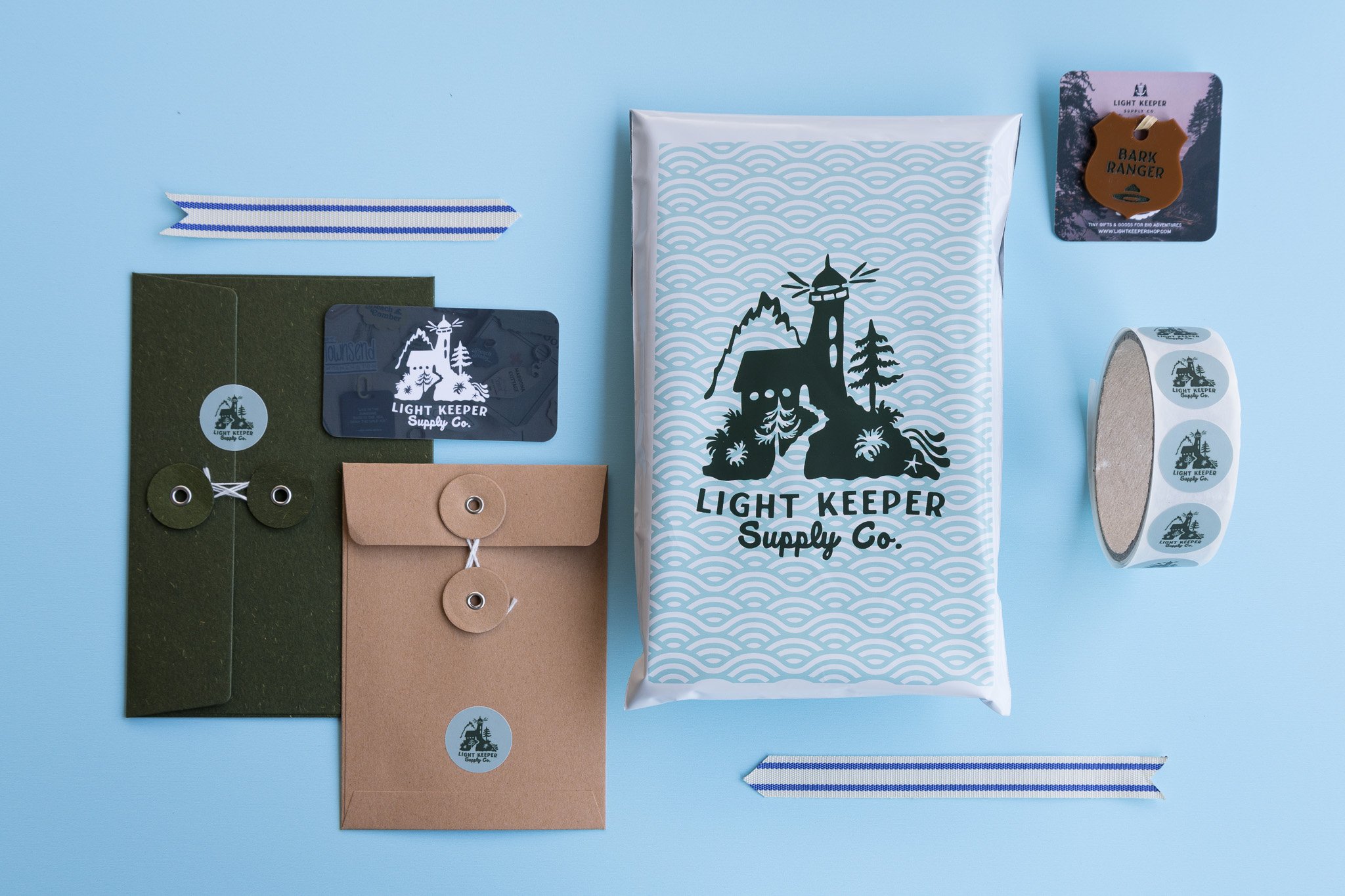 Assorted stationery and branding items from Light Keeper Supply Co., including a bar of soap, a roll of stickers, two gift envelopes, ribbon, and tags with the company's logo and lighthouse designs, all arranged on a light blue background.