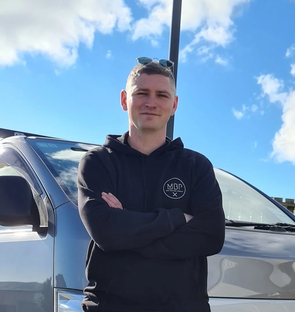 A young man with arms crossed, wearing a black hoodie with a logo, standing in front of a silver vehicle on a sunny day with blue sky and clouds.