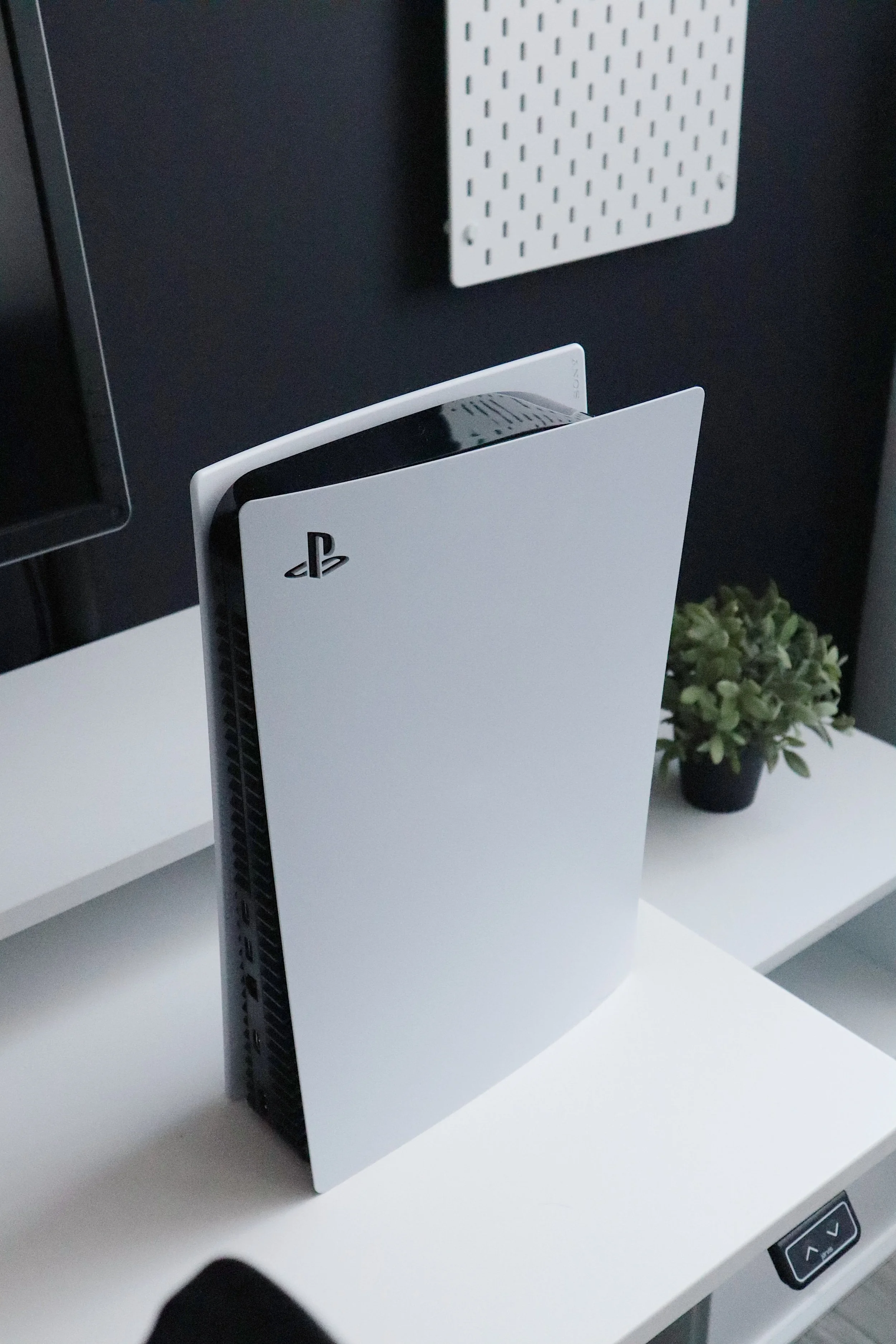 A white PlayStation 5 gaming console standing upright on a white desk with a small potted plant in the background.