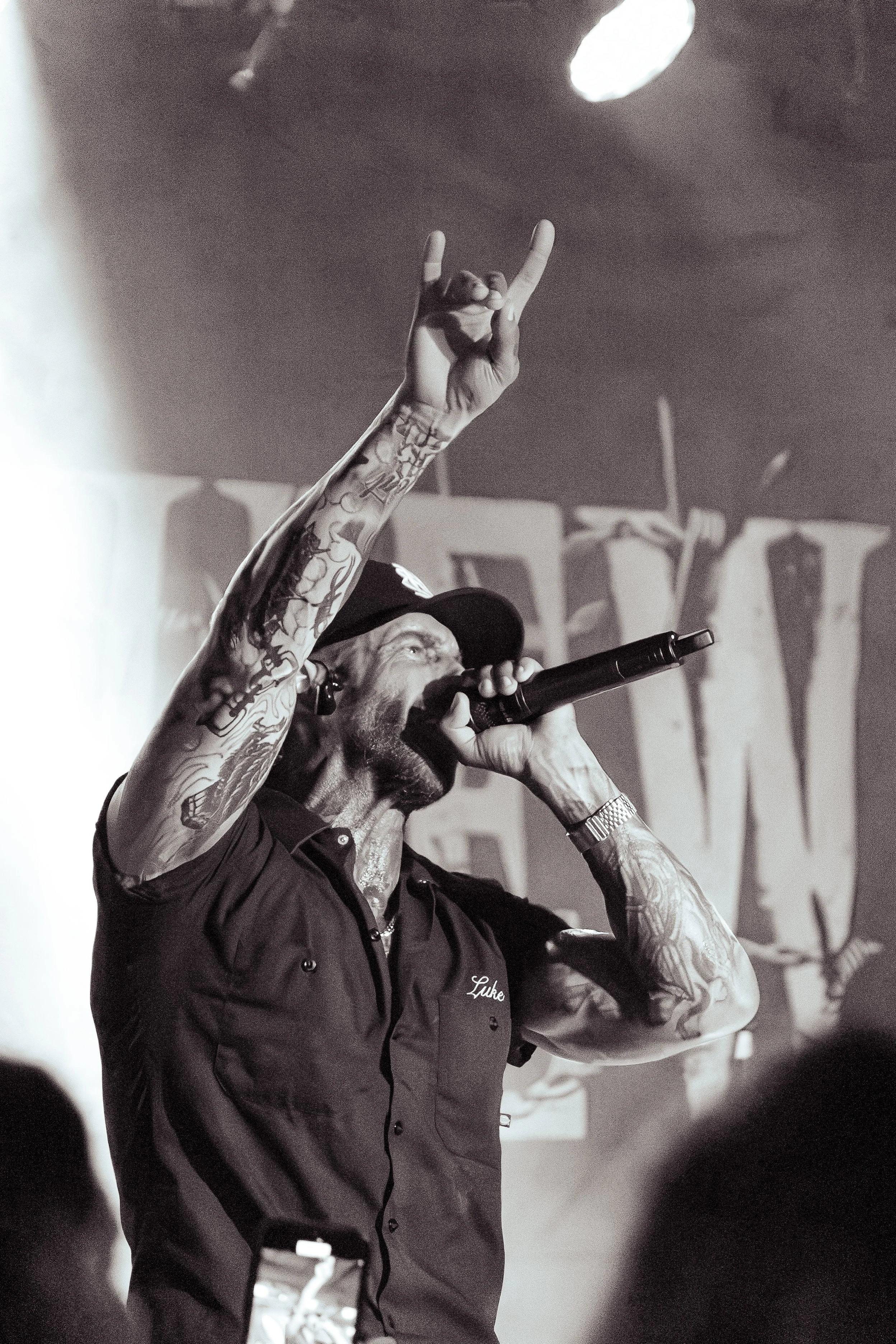 A man with tattoos on his arm singing into a microphone and making a hand gesture, wearing a shirt with the name 'Luke' on it.