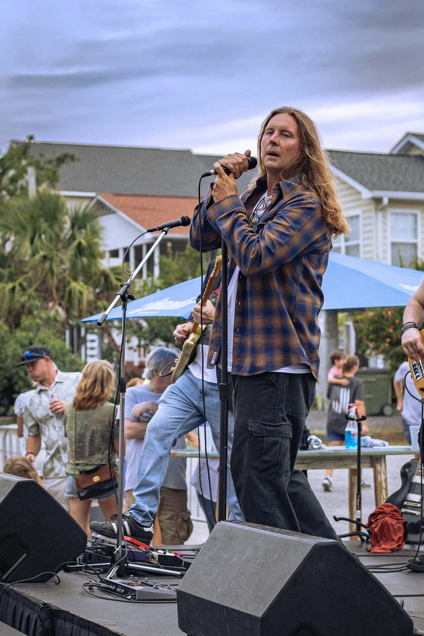 A male musician with long hair and a plaid shirt singing into a microphone on an outdoor stage during a live performance. People are gathered around, some sitting and some standing, with houses in the background under a cloudy sky.