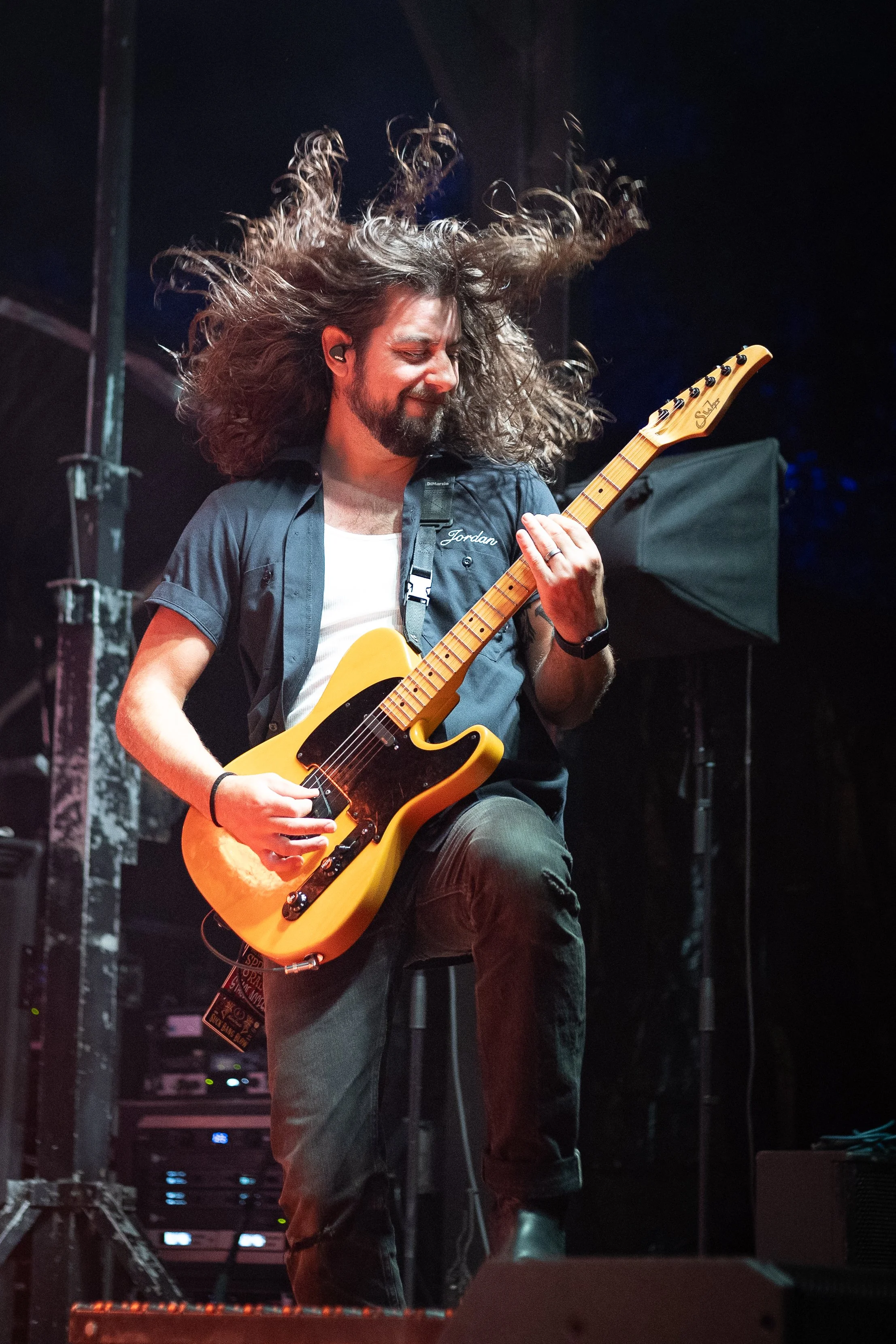 A male musician with long curly hair joyful playing an electric guitar on stage at night, wearing a black shirt with a name tag and jeans, with stage equipment and dark background visible.