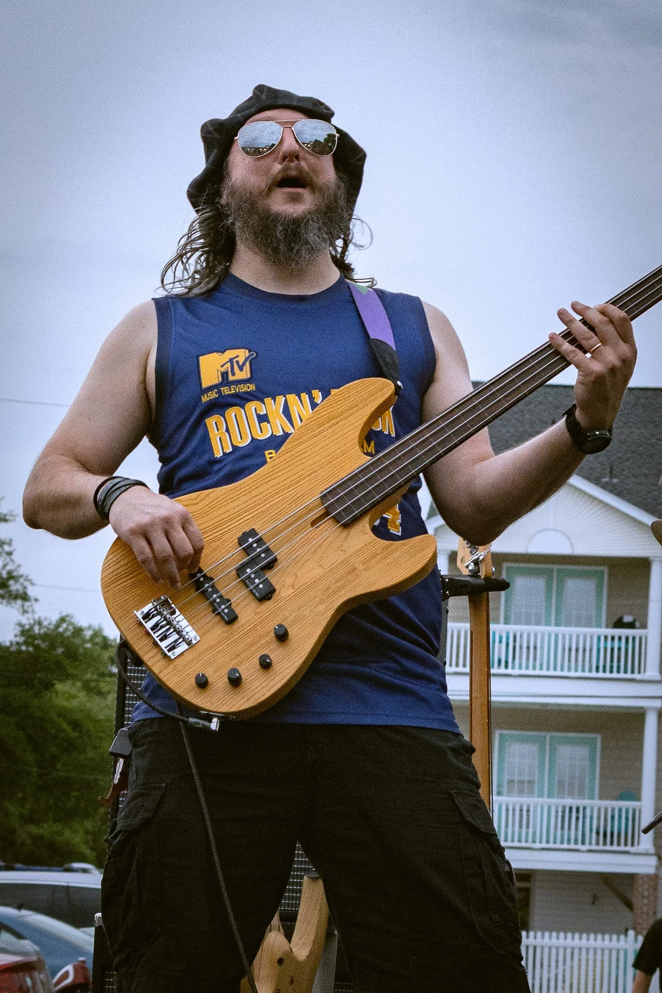 A man with a beard and sunglasses playing an electric bass guitar outdoors, wearing a sleeveless shirt and black cargo shorts.