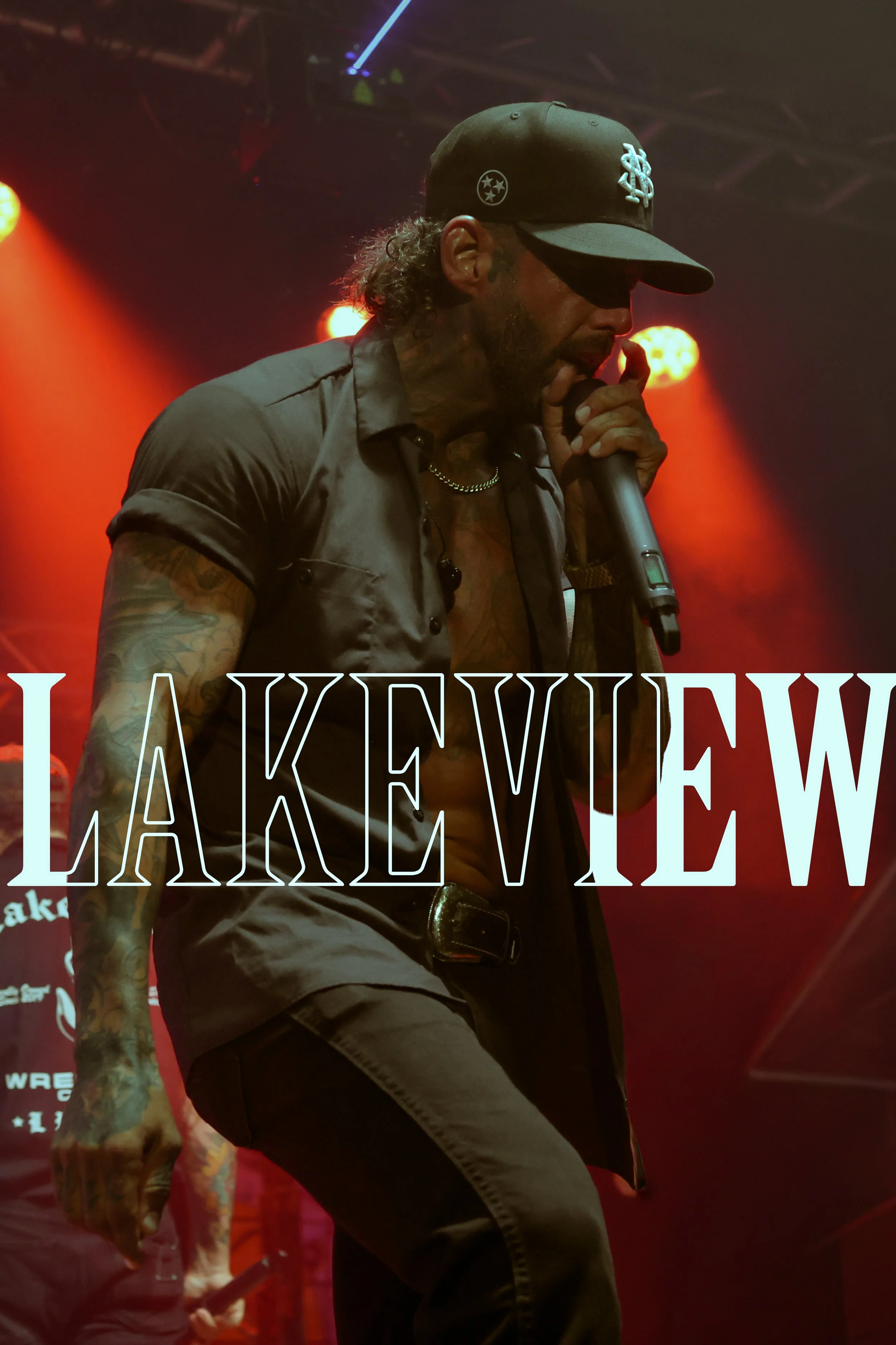 Male singer wearing a black cap and shiny gray shirt, performing on stage with red and orange lighting, and the partially visible word "LAKEVIEW" in front.