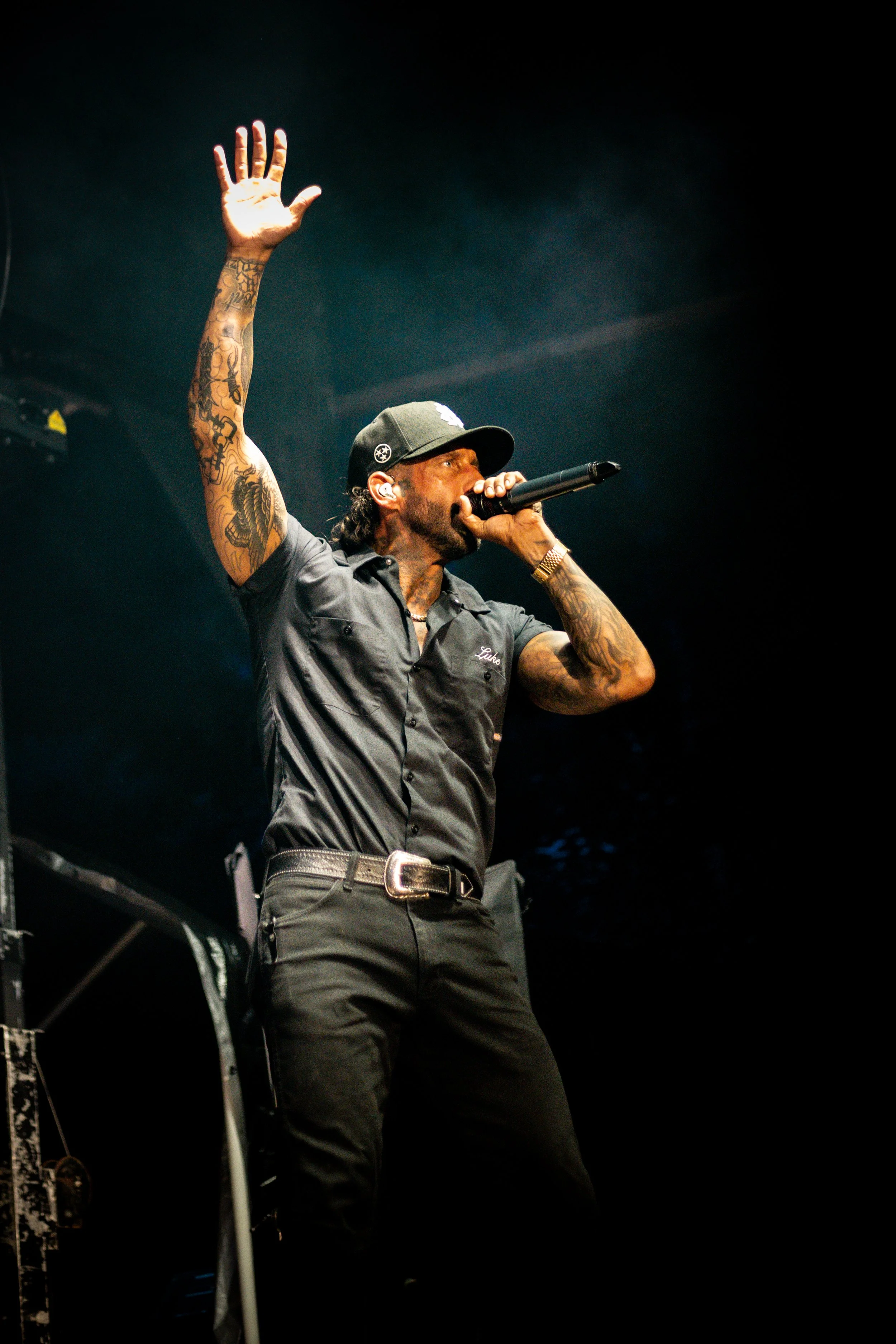 A male singer performing on stage, holding a microphone and raising one arm with an open hand, wearing a black cap, gray shirt, and black pants, with tattoos on his arm and a dark background.