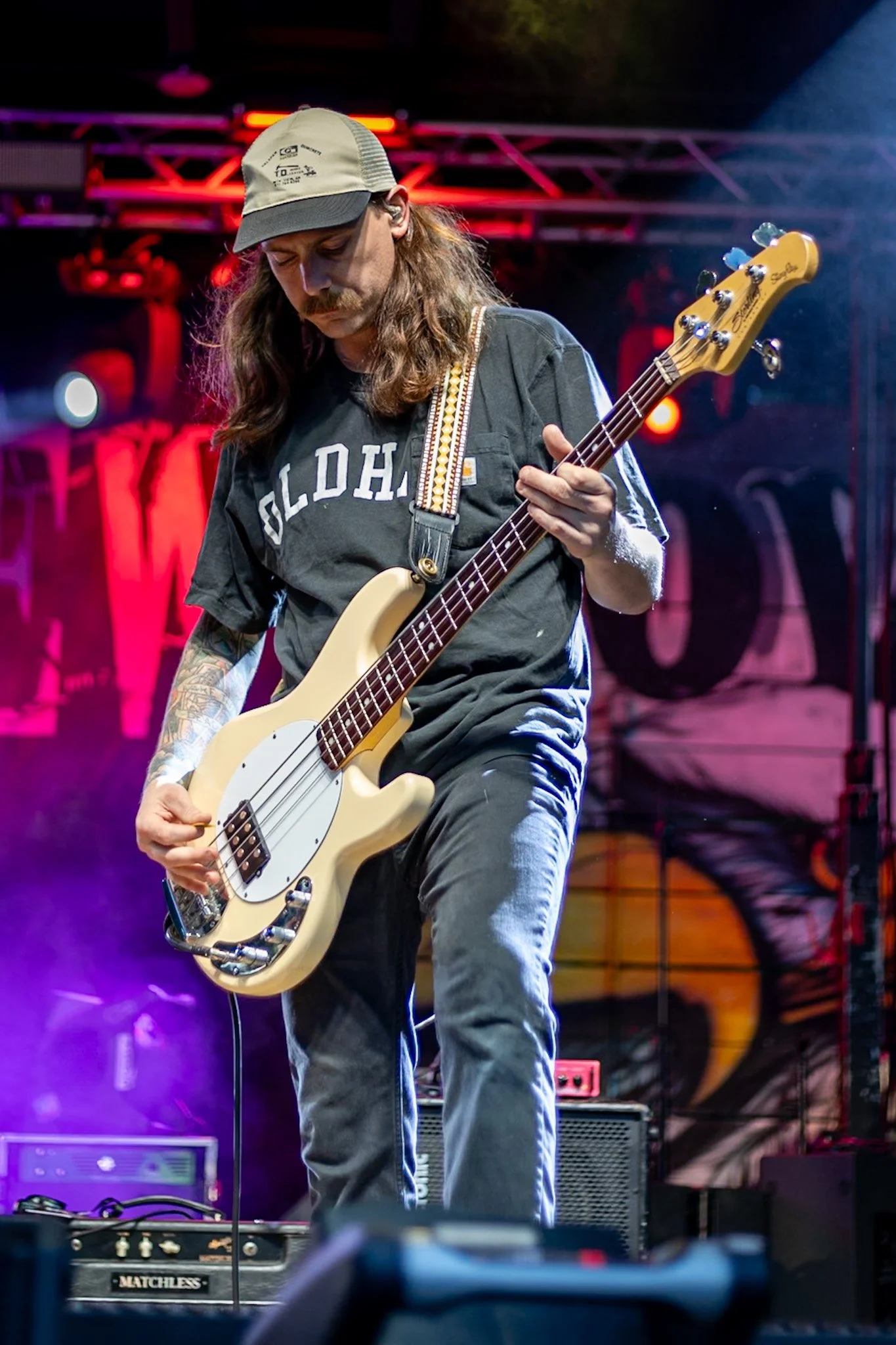 A man with long hair, a beard, and a mustache playing an electric bass guitar on stage with colorful lighting, wearing a gray cap and a black T-shirt. There are amplifiers and stage equipment in the background.