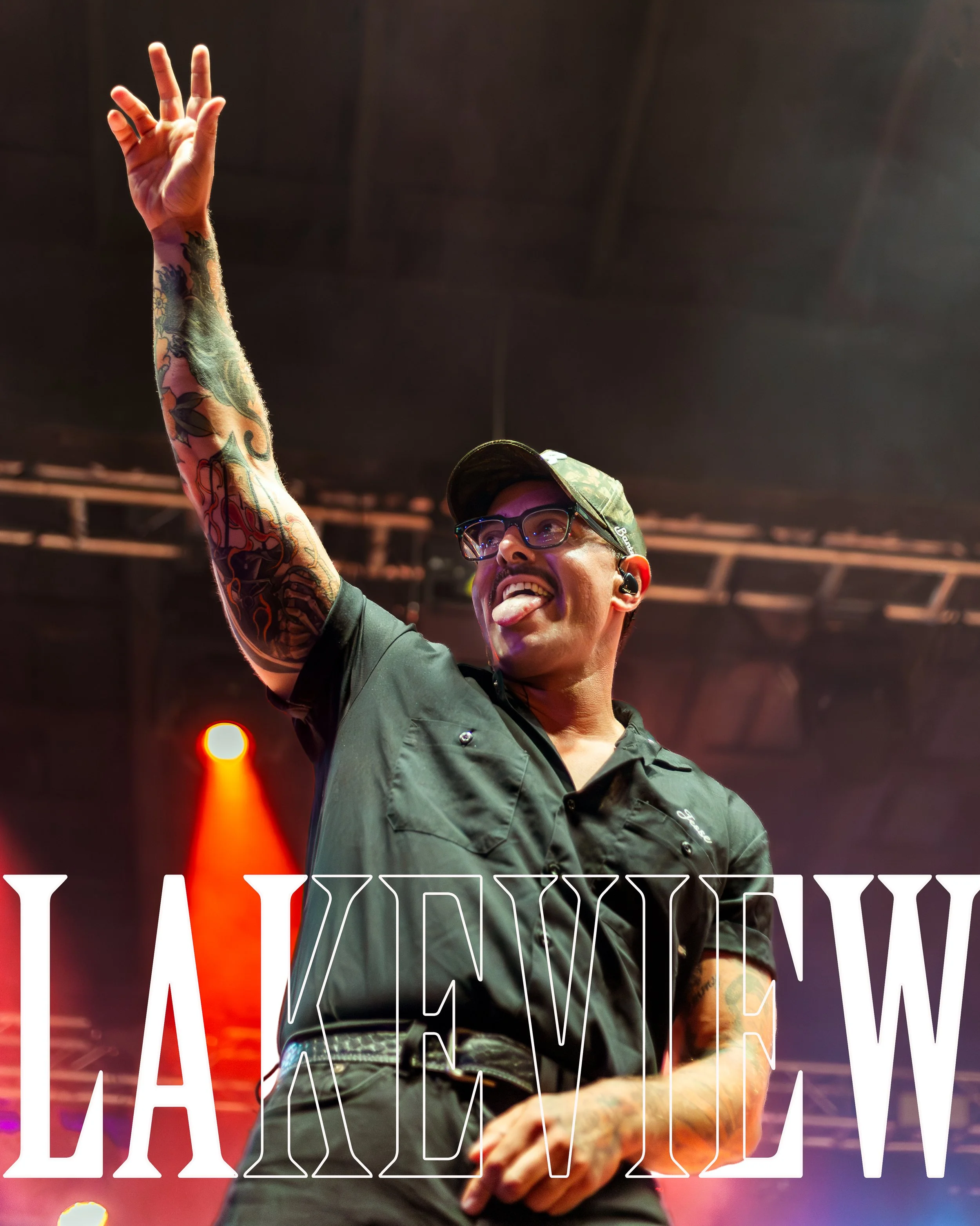 A person with tattoos on their arm, glasses, and a camouflage baseball cap is smiling and sticking out their tongue while raising one arm with fingers extended. The background shows stage lighting and a sign with partially obscured large white letter