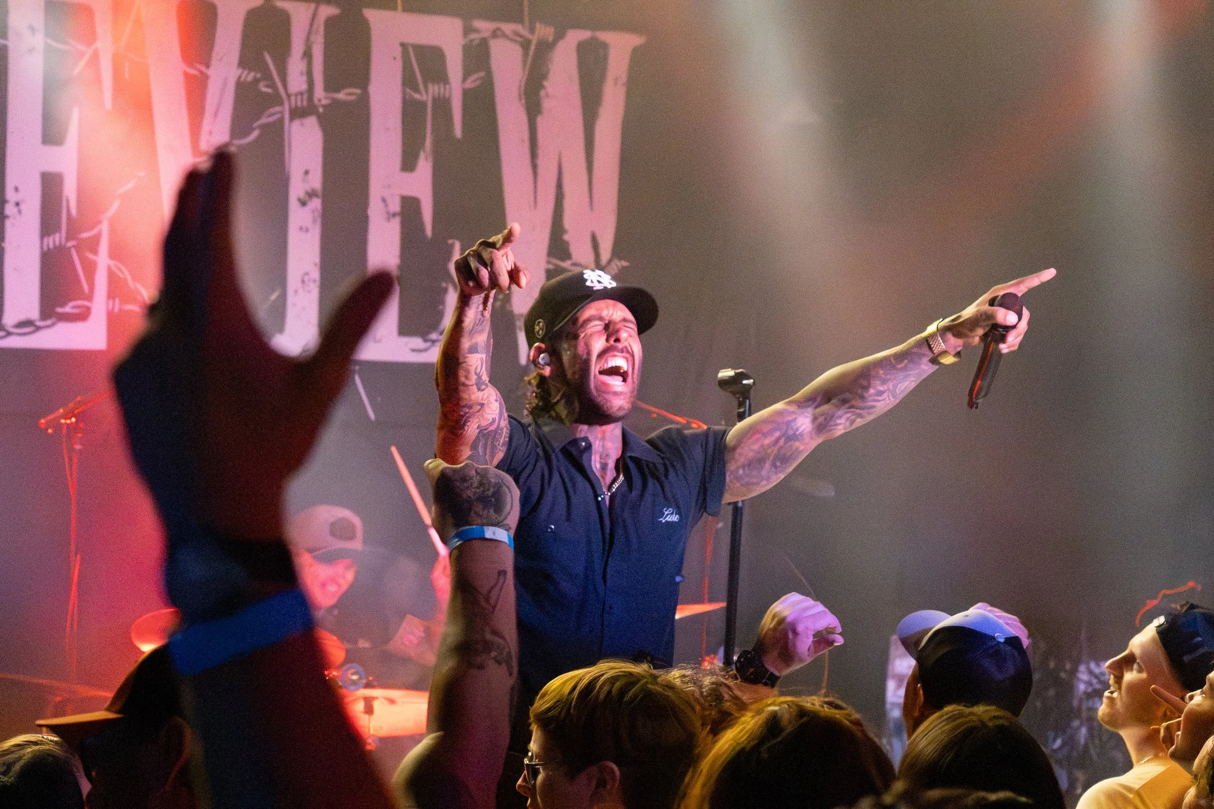 A man with tattoos, wearing a black cap and navy blue shirt, is on stage singing passionately, holding a microphone in one hand and pointing with the other. There is an audience in front of him, and a large screen behind displaying colorful abstract 