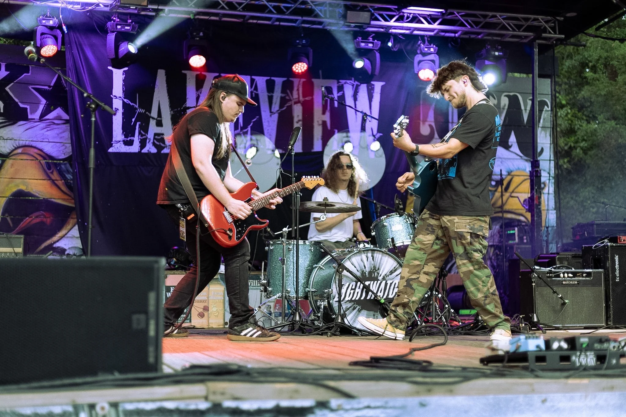 Two musicians performing on stage outdoors, one playing an electric guitar and the other playing an electric bass, with a drummer in the background. The stage has colorful lighting and a backdrop with graffiti-style art.