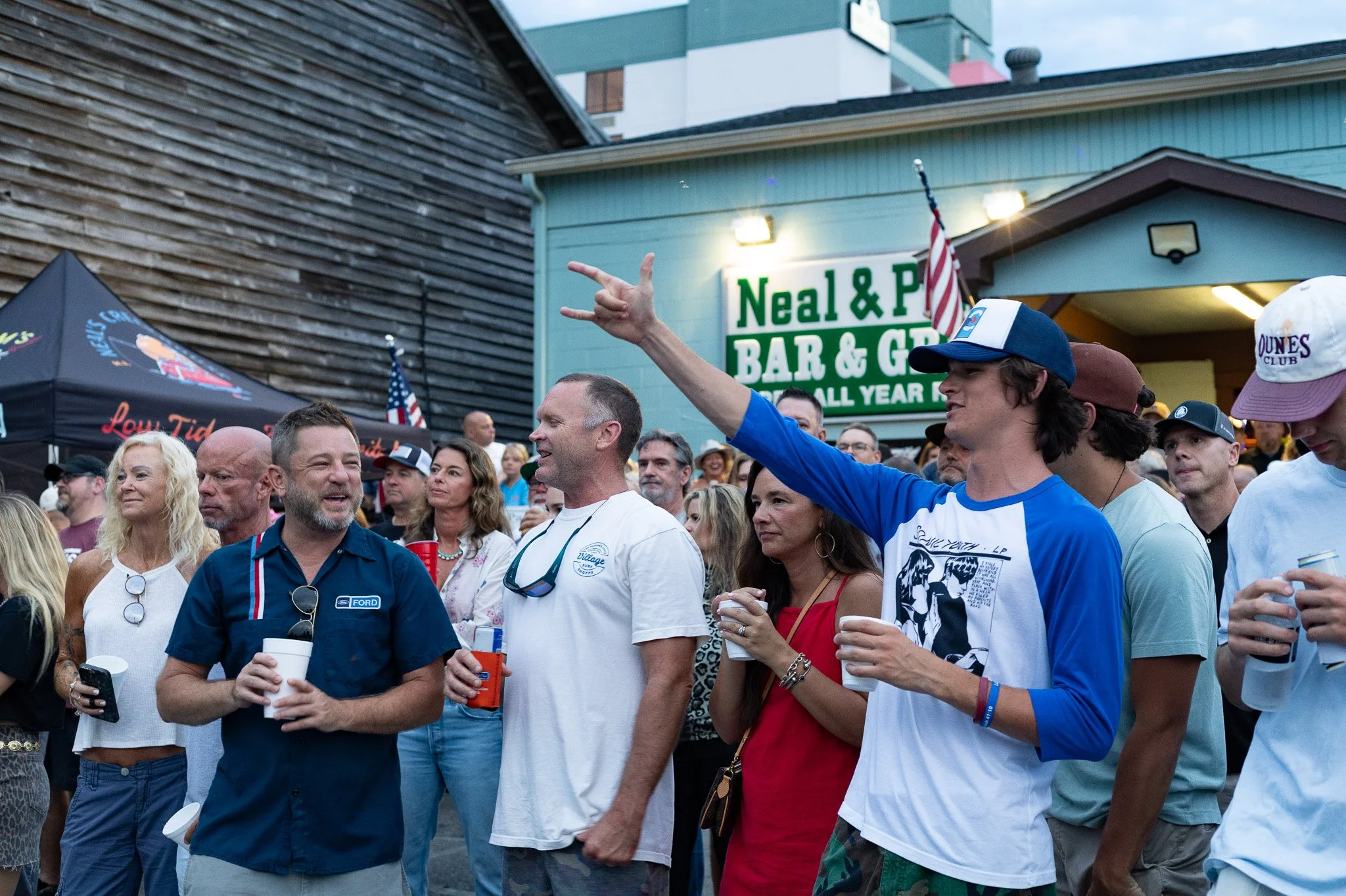 A crowd of people gathered outside Neal & P's Bar & Grill, with some individuals holding drinks, engaging in conversations, and wearing casual summer clothing. A man in a blue shirt and a woman in a red dress are notable in the foreground.