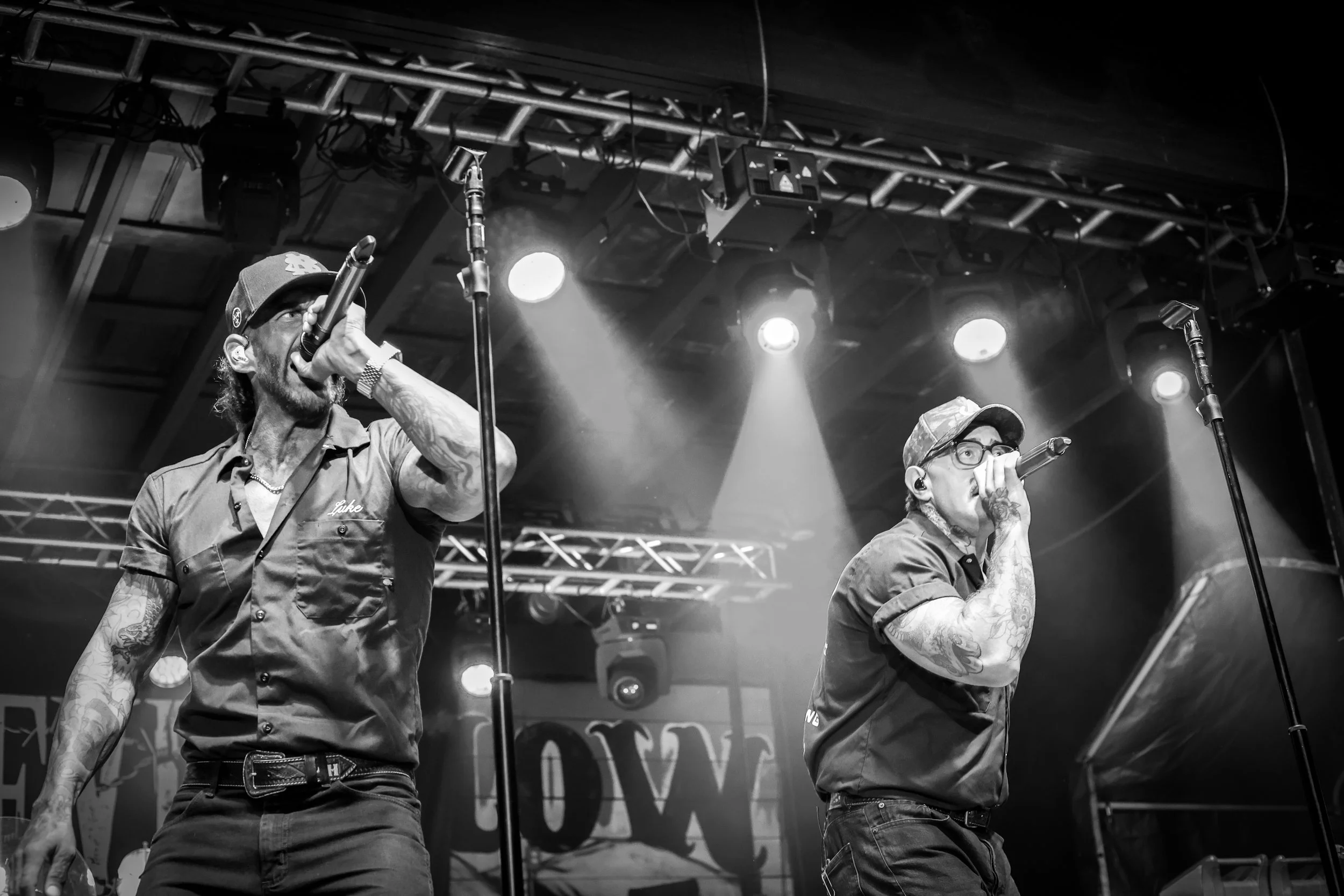 Two male performers singing into microphones on stage during a concert, illuminated by stage lights, with visible stage equipment and scaffolding overhead.